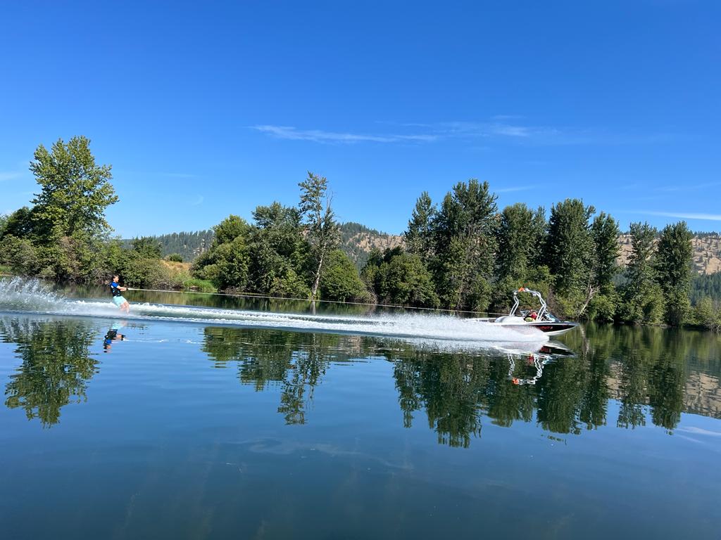 A skier on calm, glassy water behind a boat, hills reflected in the lake